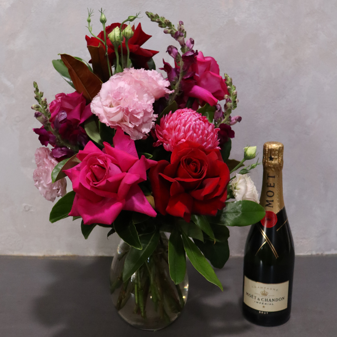 The Sparkling Charm Flower Gift Set by Flowers on Norton St features a bouquet of pink, red, and magenta passion flowers with roses and carnations in a glass vase, paired with Moët & Chandon champagne. Ideal for Valentine's Day, it beautifully displays lush green foliage on a gray surface.