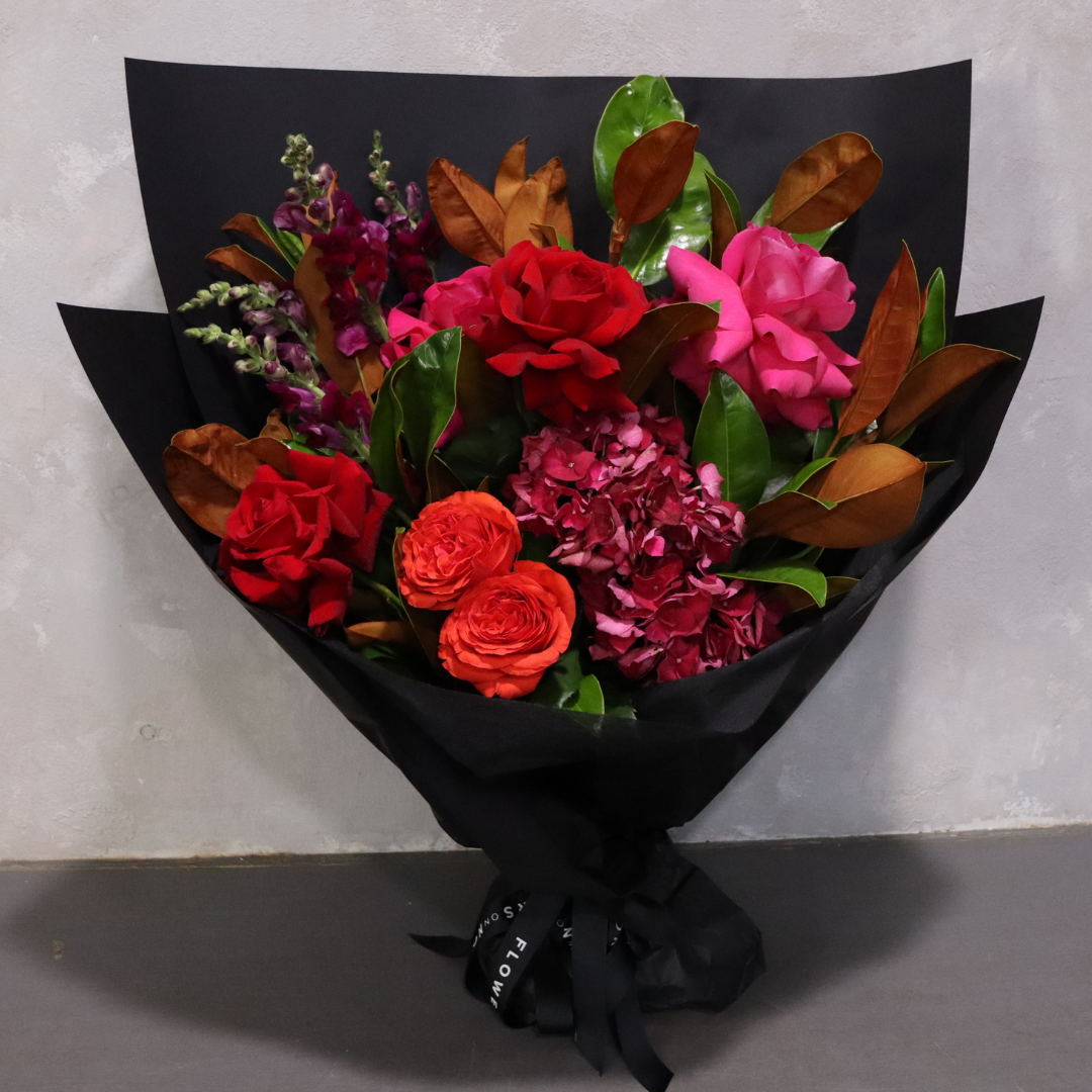 The Utterly Entranced Red Bouquet by Flowers on Norton St, from the Valentine's Day Collection, features red and pink roses with vibrant orange blooms and pink hydrangeas, accented by greenery. It's elegantly wrapped in black paper with a matching ribbon set against a simple gray backdrop.