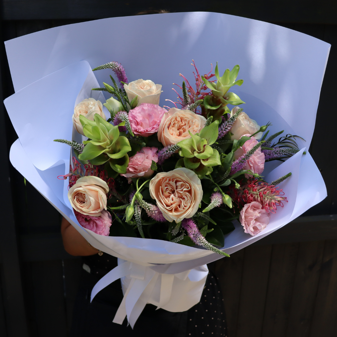 The Summer Crush | January Mixed Bouquet by Flowers on Norton St showcases peach roses, pink carnations, and Siam Tulips with green accents in an elegant white paper wrap with a white ribbon, set against a dark fence backdrop.