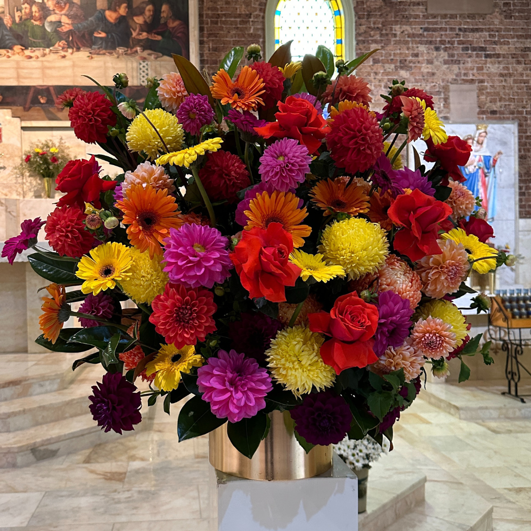 The Large Gold Pot Arrangement by Flowers on Norton St presents a radiant collection of red, yellow, purple, and orange flowers. This impressive seasonal display creates a striking contrast against the stained glass windows and religious paintings in the background.