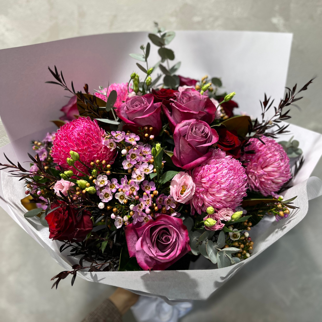 A bouquet known as the "Berry Bliss Purple Bouquet" by Flowers on Norton St, showcasing pink roses, vibrant pink chrysanthemums, red roses, and small pink blossoms, arranged with green foliage and encased in white paper.