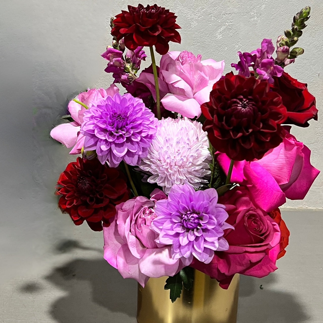 The Passion Pop Purple Bouquet by FlowersonNortonSt features a gold pot brimming with flipped roses, burgundy dahlias, and purple chrysanthemums against a plain gray background. The arrangement is colorful and lush.