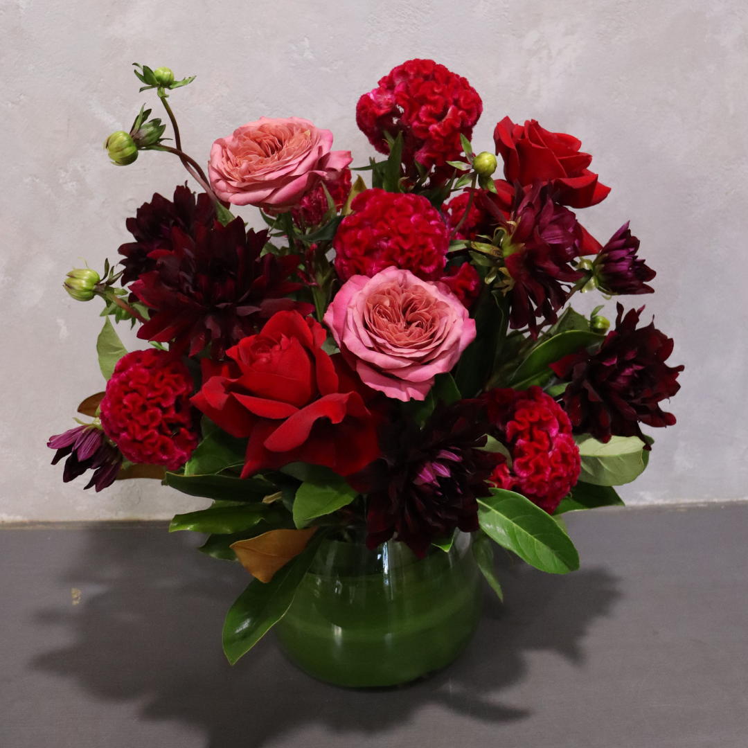 The Pure Passion Mixed Bouquet by Flowers on Norton St features a captivating mix of red and pink roses, dark red dahlias, green leaves, and small buds in a green vase. This floral delight against a light gray backdrop creates an aura of romantic bliss.
