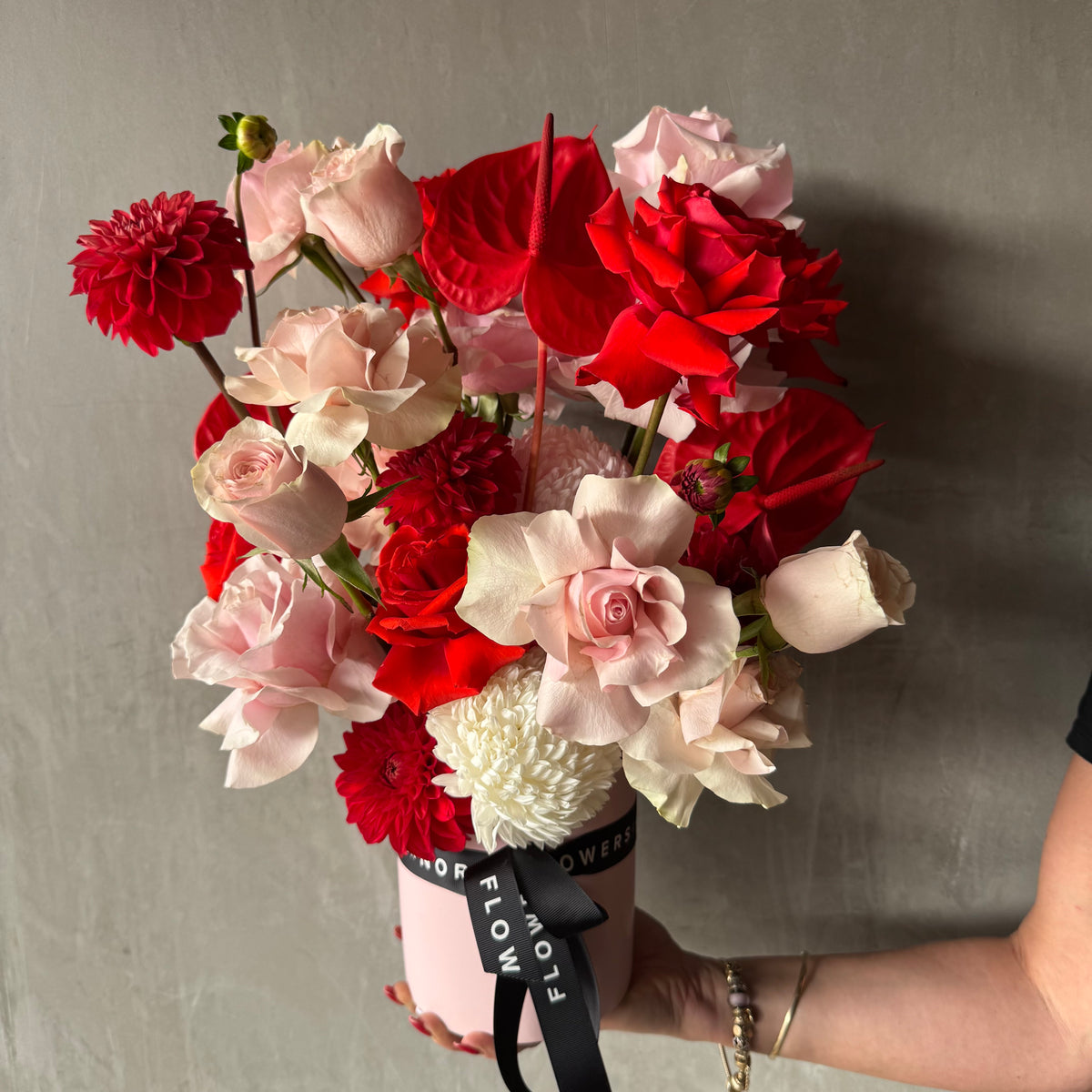 A hand displays the Be My Valentine Vase Arrangement by Flowers on Norton St, featuring Colombian red and pink roses, dahlias, anthuriums, and more in a pink pot with a black ribbon against a gray backgroundāperfect for Valentine's Day.