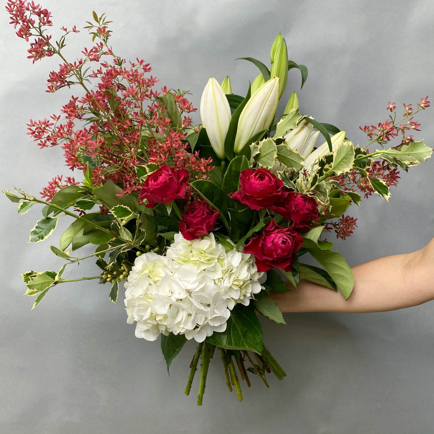 The Christmas Mixed Flower Bouquet by Flowers on Norton St, held against a gray backdrop, showcases white lilies, hydrangeas, red roses, and red berries with lush greenery. An arm presents this vibrant arrangement as the ideal accent for your holiday décor.