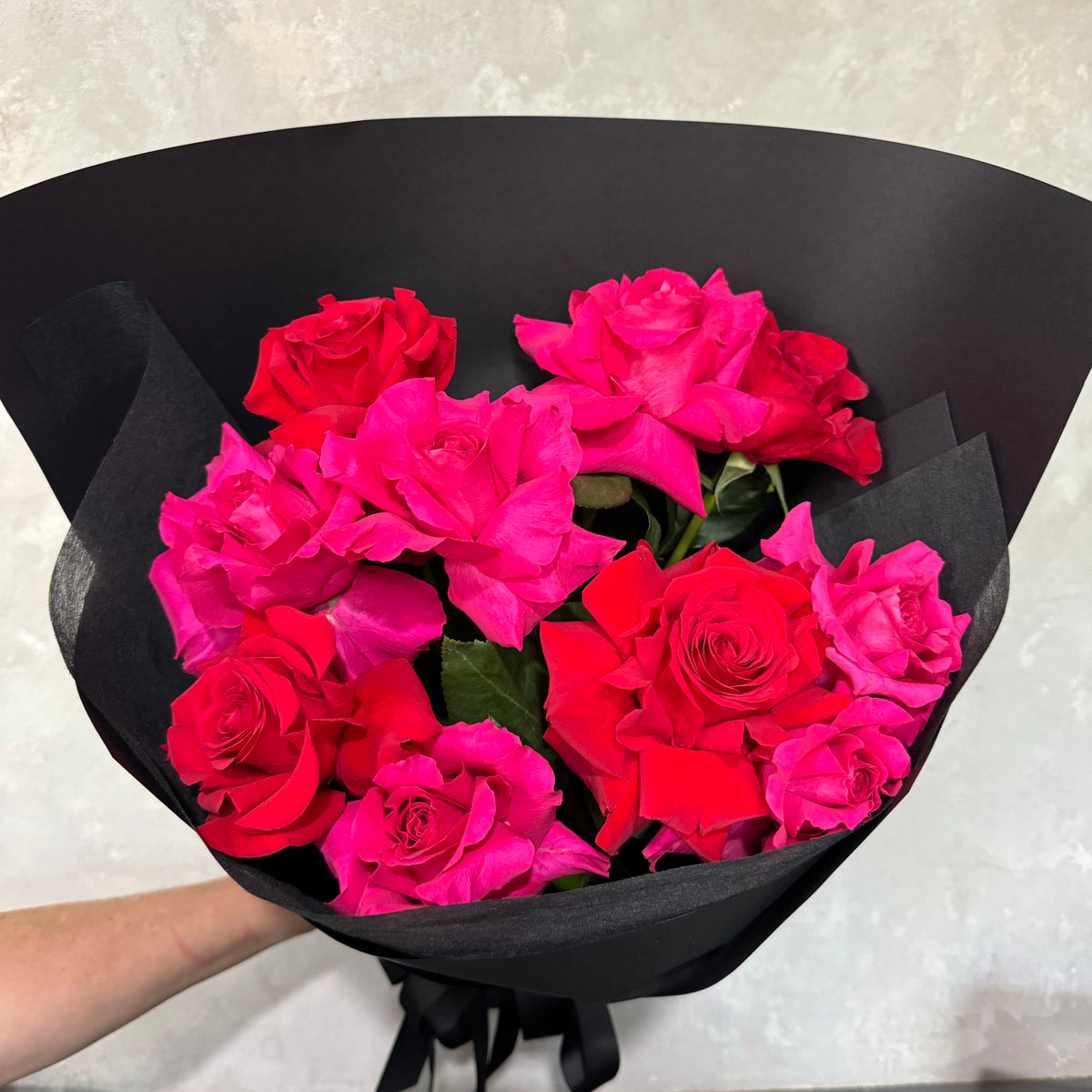 The Pretty like a Primrose February Pink Bouquet by Flowers on Norton St features vibrant Colombian hot pink and red roses elegantly wrapped in sleek black paper against a light gray background.