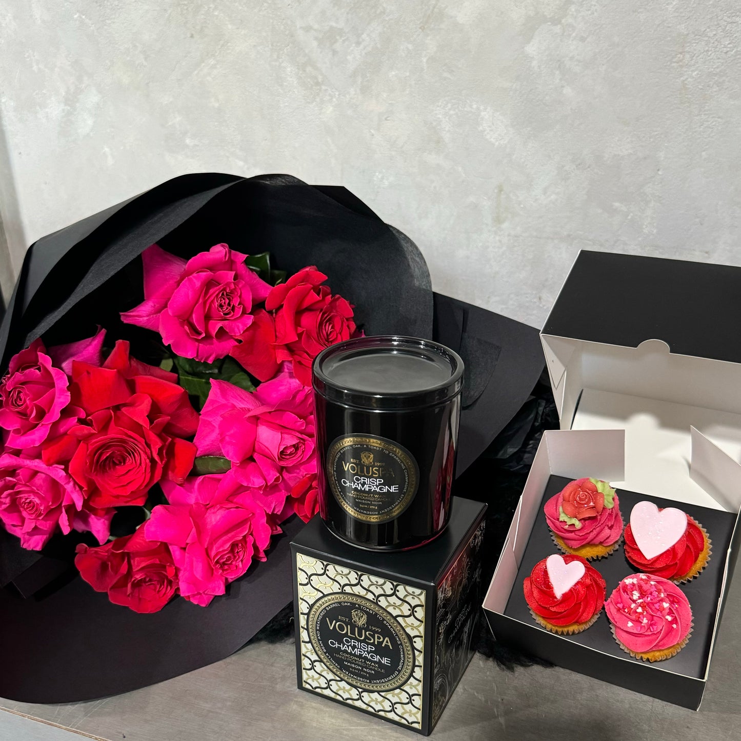 The Valentine's Rose Bouquet + Cupcakes from Flowers on Norton St features romantic pink roses in black paper with a VOLUSPA Champagne black candle, accompanied by a sleek box of three Valentine's Day cupcakes adorned with pink frosting and heart-shaped toppings.