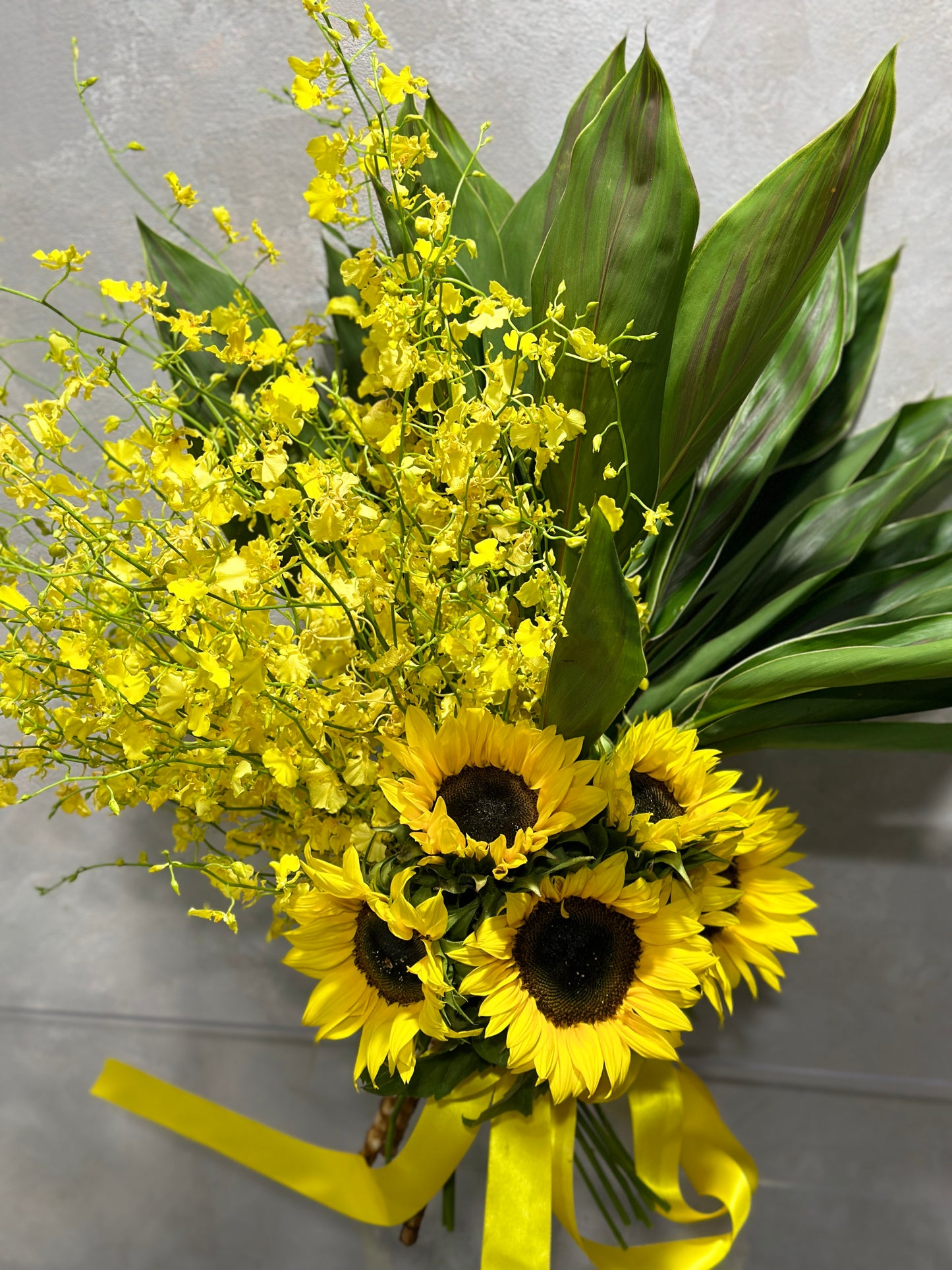 Discover the Radiant Rays Bouquet from Flowers on Norton St, featuring vibrant sunflowers and striking yellow dancing lady orchids, elegantly accented with lush green leaves. The stems are beautifully tied with a yellow ribbon and displayed against a gray background.
