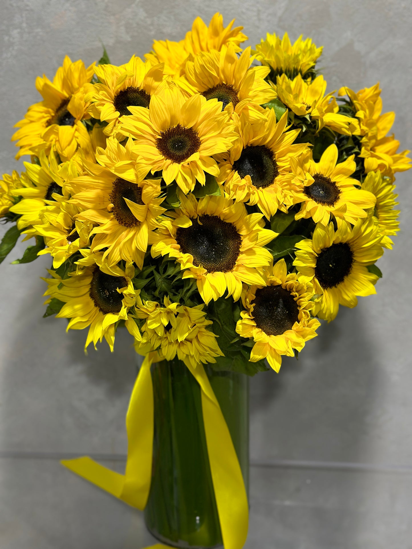 The Flowers on Norton St "Spectacular Sunnies Vase" features a striking arrangement of vibrant yellow sunflowers with dark centers, beautifully displayed in a tall glass vase. Accentuated by a yellow ribbon, this natural beauty stands out against a neutral gray background.