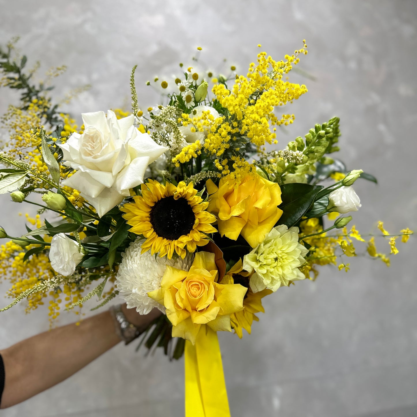 A hand holds the vibrant Lemon Fizz bouquet from Flowers on Norton St, showcasing sunflowers, yellow and white roses, and an array of chrysanthemums against a neutral background. The stems are elegantly tied with a yellow ribbon.