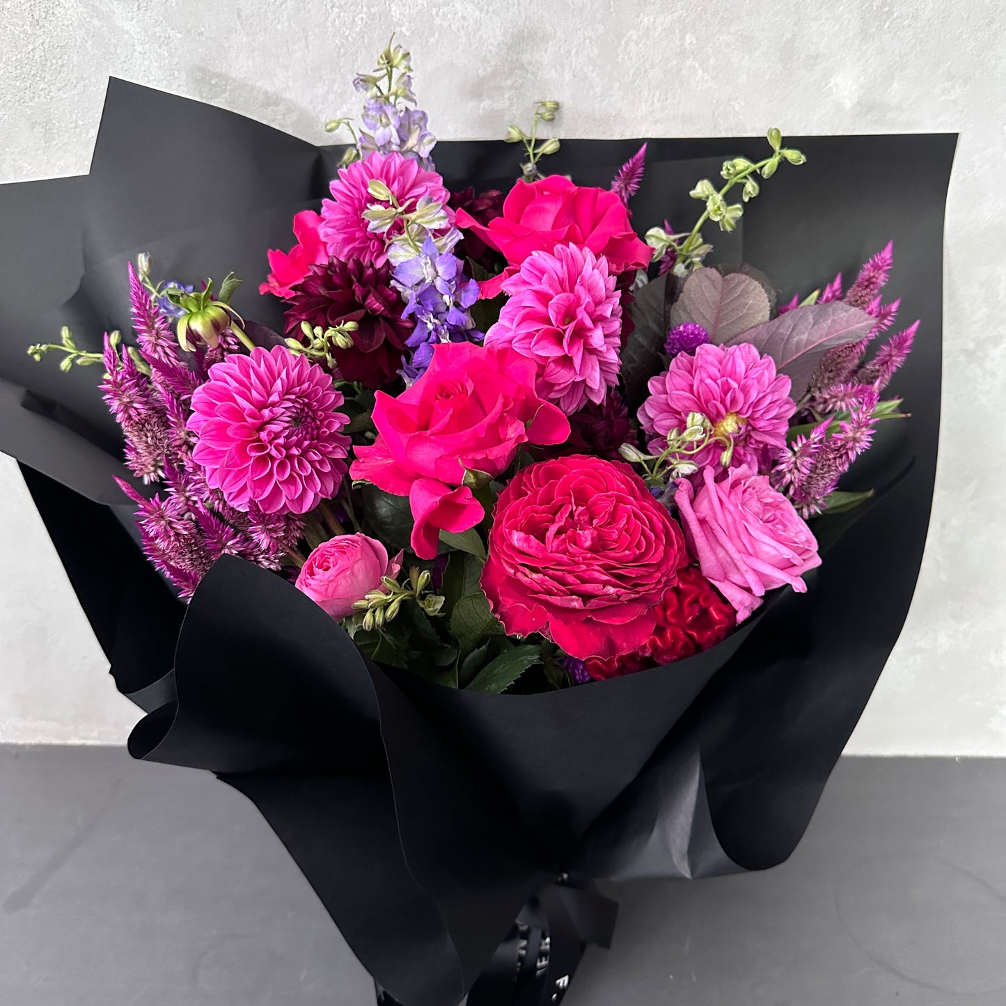 Introducing "The $109 Bouquet" by Flowers on Norton St: a budget-friendly floral arrangement showcasing vibrant hot pink and purple seasonal blossoms, such as dahlias and roses. Wrapped in sophisticated black paper, this bouquet is beautifully presented against a light gray backdrop.