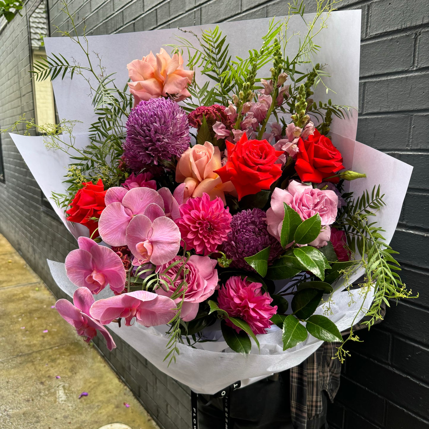 The Bejewelled Mixed Flower Bouquet by Flowers on Norton St showcases a vibrant assortment of red roses, pink orchids, and dahlias, complemented by various purple and pink flowers. Accented with greenery and elegantly wrapped in white paper, this beautiful arrangement is displayed against a brick wall.