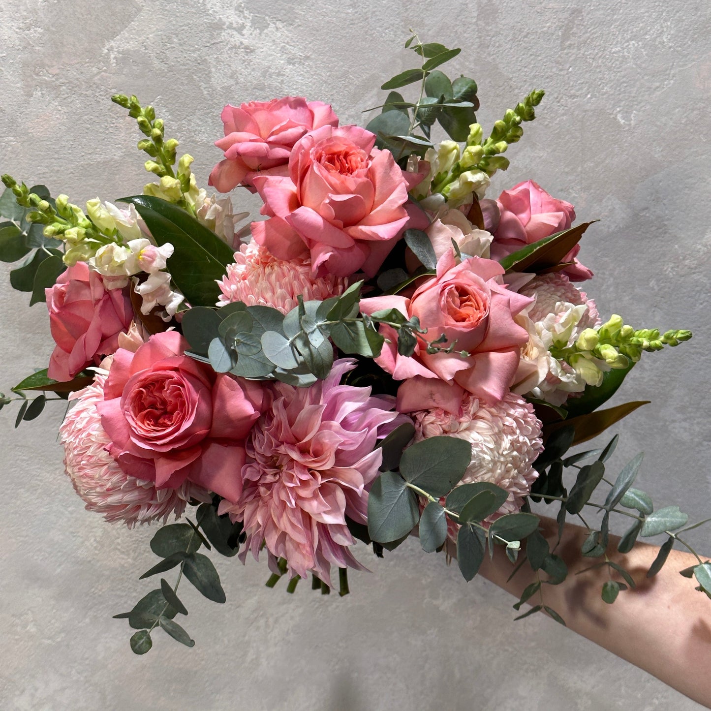 A hand holds the "Pretty in Pink Mixed Flowers" bouquet by Flowers on Norton St, featuring a seasonal mix of pink roses, dahlias, and snapdragons accented with eucalyptus leaves, against a gray background.