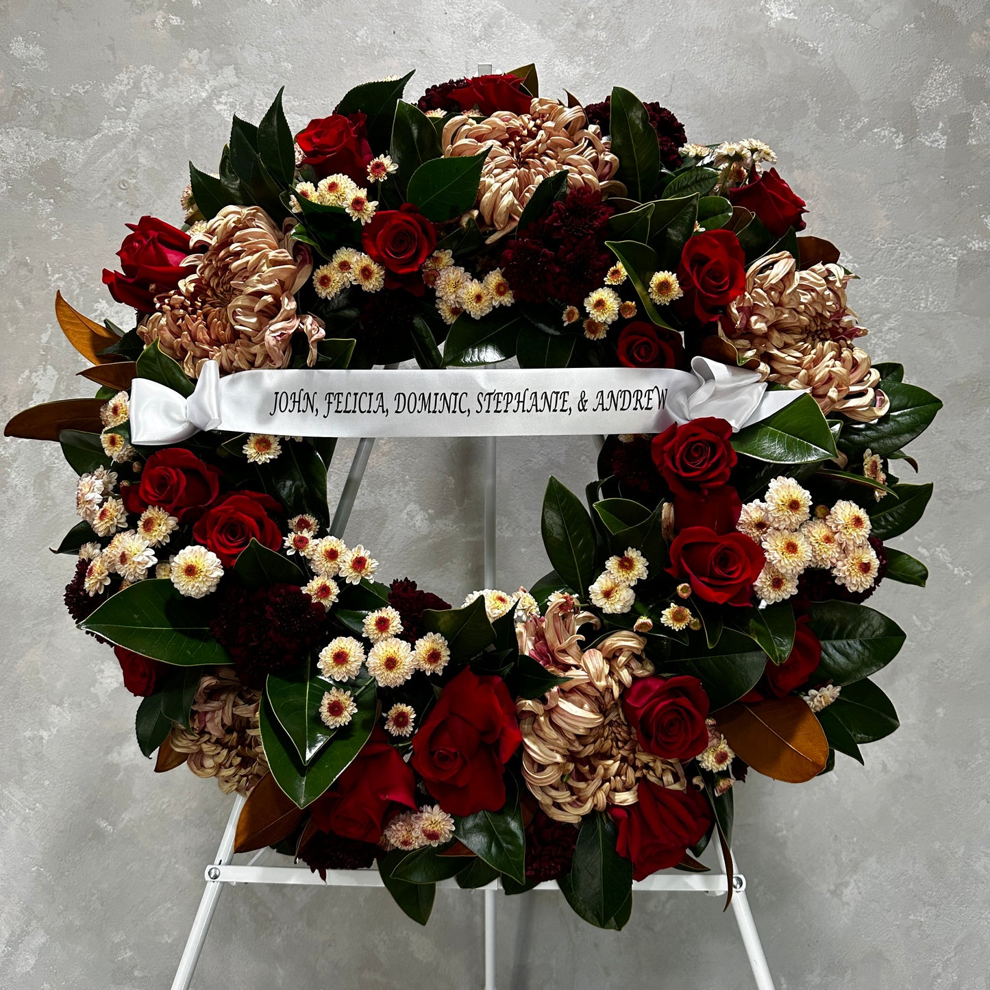 The Condolences Wreath by FlowersonNortonSt is a stunning arrangement featuring red roses, white and pink blooms, and lush green foliage. A custom printed ribbon displays the names John, Felicia, Dominic, Stephanie, & Andrew. The wreath is elegantly presented on a white easel against a gray backdrop.