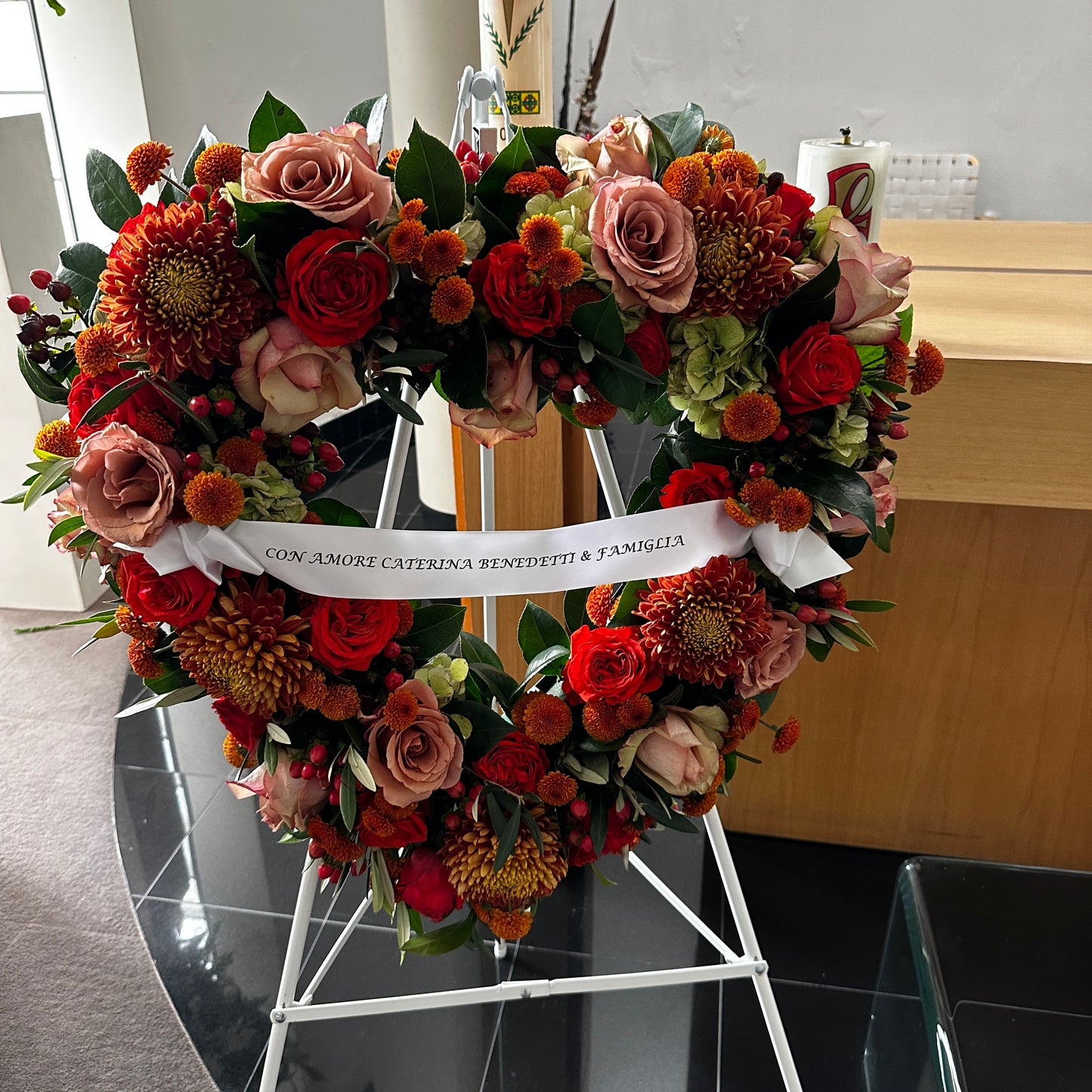 The Sympathy Heart Wreath by FlowersonNortonSt features premium red, orange, and pink flowers with a white ribbon inscribed in Italian. Displayed elegantly on a white stand in a room with dark tiled floors and a wooden counter.