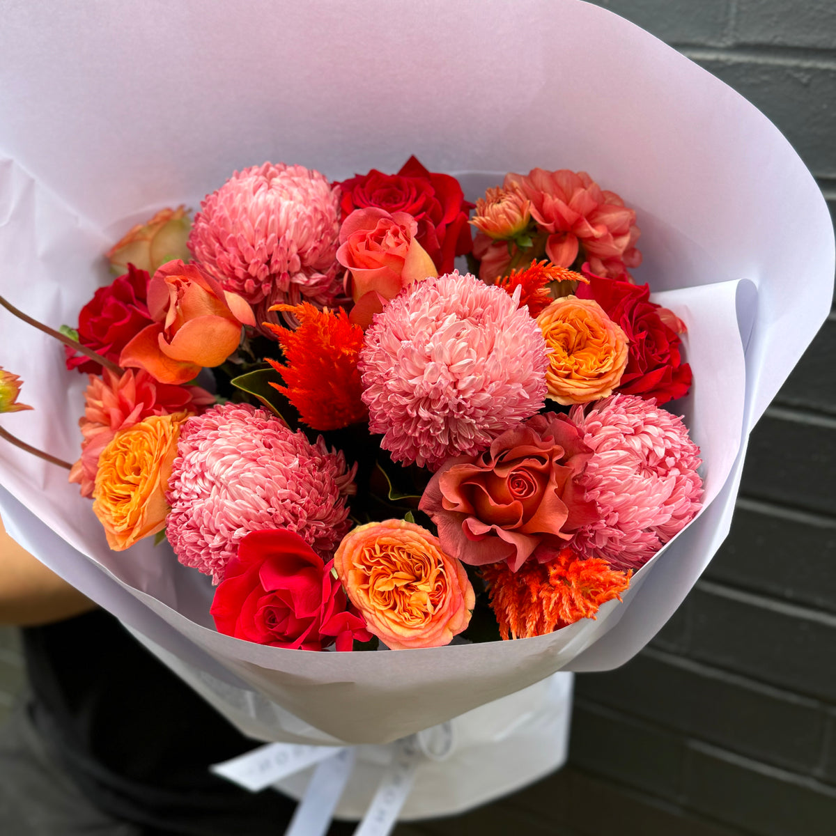 An Autumn Flower Bouquet by Flowers on Norton St showcases a stunning array of pink, red, orange, and peach chrysanthemums and roses, elegantly wrapped in white paper and set against a dark background.