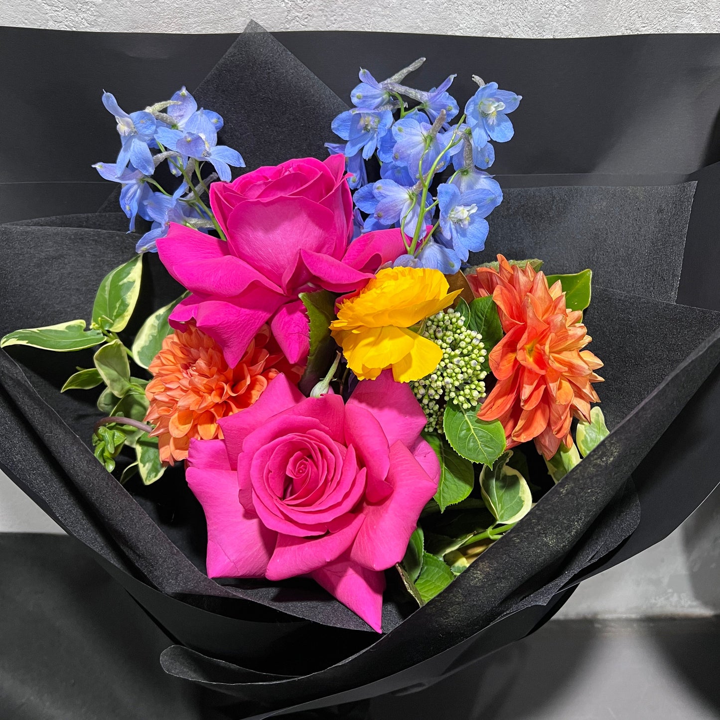 Introducing the "Posies of Joy" by FlowersonNortonSt – a stunning bouquet featuring pink roses, complemented by vibrant orange and yellow flowers, and delicate blue blossoms. Arranged by skilled florists with lush green leaves, it is elegantly wrapped in black paper for a striking contrast against the colorful blooms.