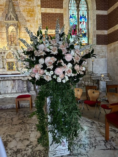 The Deluxe Waterfall Flower Arrangement by Flowers on Norton St, featuring pink and white florals with lush green foliage, graces the church, enhancing the stone altar, stained glass windows, and wooden chairs.