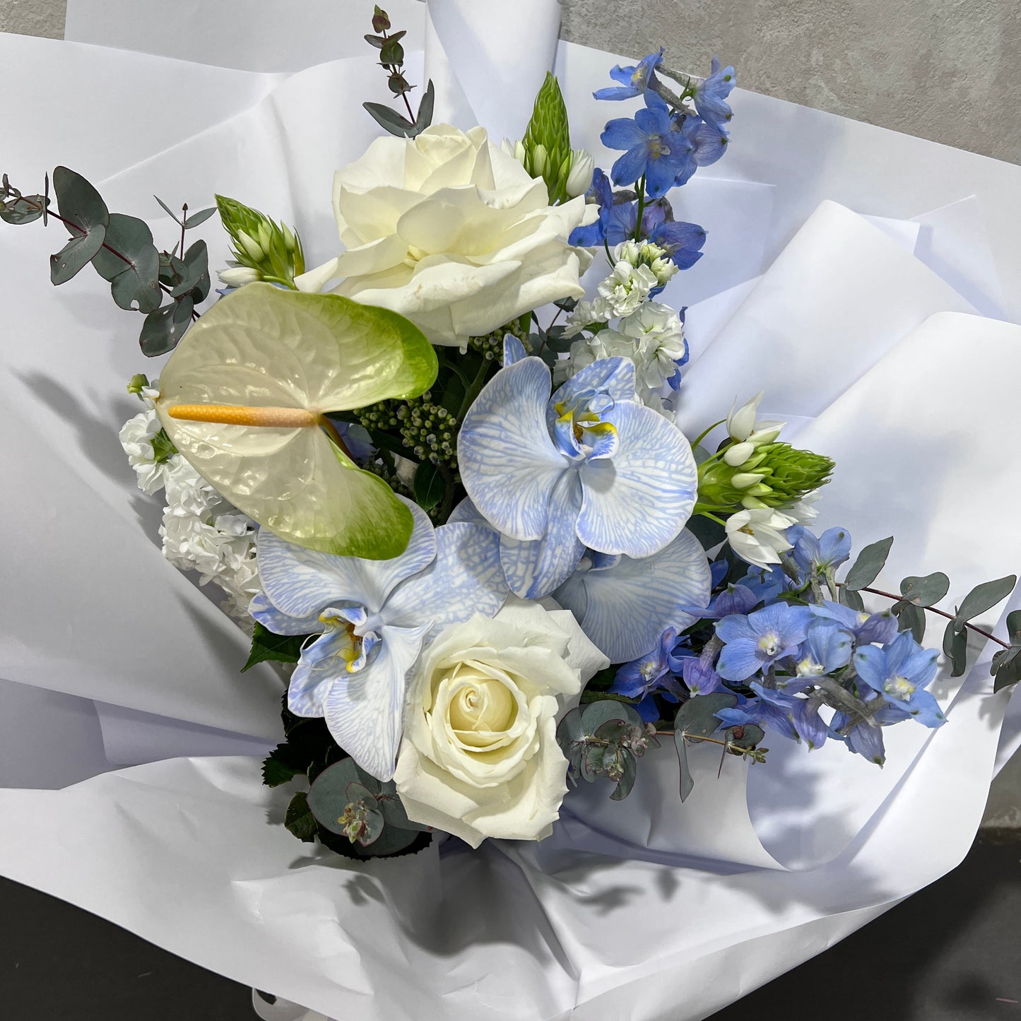 Introducing the Blue Daze Seasonal Bouquet by FlowersonNortonSt, showcasing a captivating array of white and pale blue blooms including roses, Blue Daze phalaenopsis orchids, and anthuriums. This elegant bouquet is beautifully accented with green leaves and eucalyptus, all wrapped in crisp white paper.
