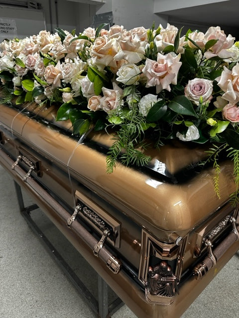 Displayed indoors is an American Style Domed Lid Casket Cover by Flowers on Norton St, adorned with premium flowers, featuring an arrangement of white and pale pink roses accented by lush green leaves.