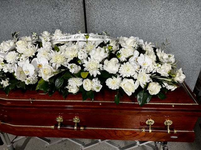 The Double Raised Lid Flower Casket Cover by Flowers on Norton St features premium flowers such as white roses and lilies. A message ribbon drapes over the blooms, and the casket is placed on a stand against a plain backdrop.