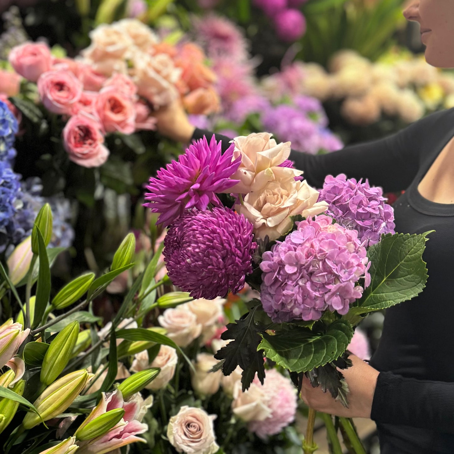 A person holds a vibrant "Florist Choice Bouquet" from Flowers on Norton St, featuring pink and purple chrysanthemums, hydrangeas, and pale pink roses, surrounded by more flowers in the lush garden.