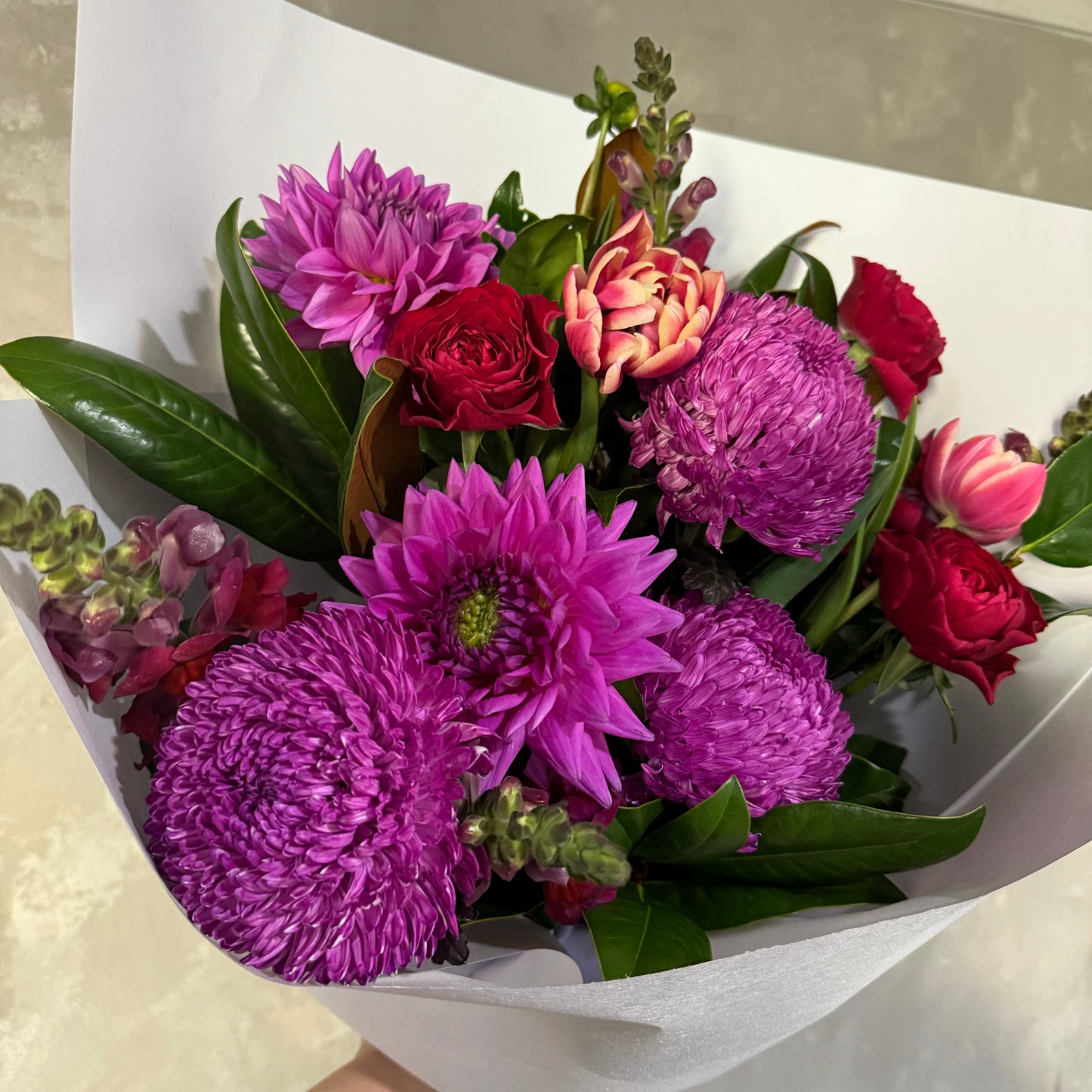 Introducing "The $89 Bouquet" by Flowers on Norton St—a vibrant arrangement of seasonal flowers that includes purple chrysanthemums, pink dahlias, red roses, and lush green leaves. Elegantly wrapped in white paper, this bouquet is perfect for any special occasion.