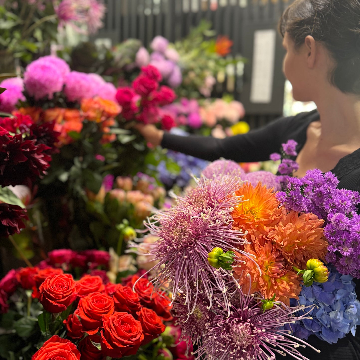In a bustling floral shop, a person with short dark hair holds Flowers on Norton St's Florist Choice Bouquet. Bursting with purple, orange, and pink blooms among vibrant arrangements, they reach for more pink flowersāideal for affordable delivery to brighten any day.