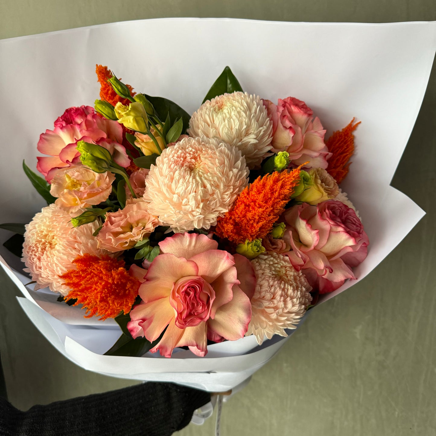 The Warm Embrace bouquet by Flowers on Norton St features an elegant blend of pink peonies, orange cockscomb, and white chrysanthemums, all beautifully wrapped in white paper. Ideal for Mother's Day, this exquisite arrangement is set against a serene muted green background.