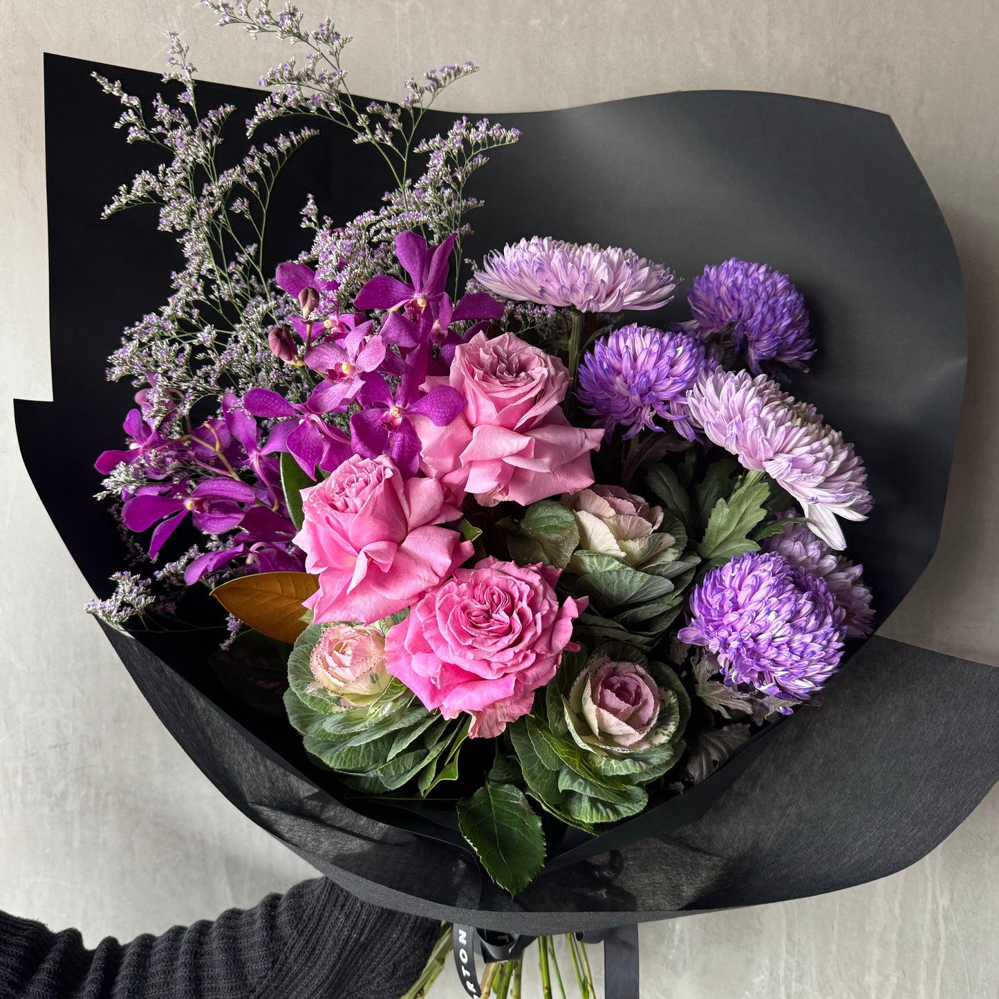 Introducing "A Mother's Love Purple Bouquet" by Flowers on Norton St: an exquisite floral arrangement that includes pink roses, purple asters, and delicate lavender blooms interwoven with lush green leaves, all elegantly wrapped in black paper. This stunning bouquet is the perfect way to celebrate the beauty and grace of Mother's Day.
