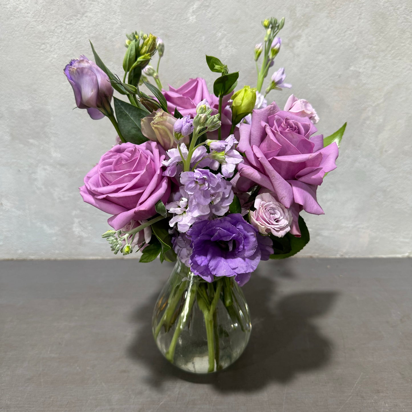 A Viola Vintage Vase from Flowers on Norton St beautifully displays a charming arrangement of purple and lavender roses, eustomas, and small white seasonal flowers against a simple gray backdrop.