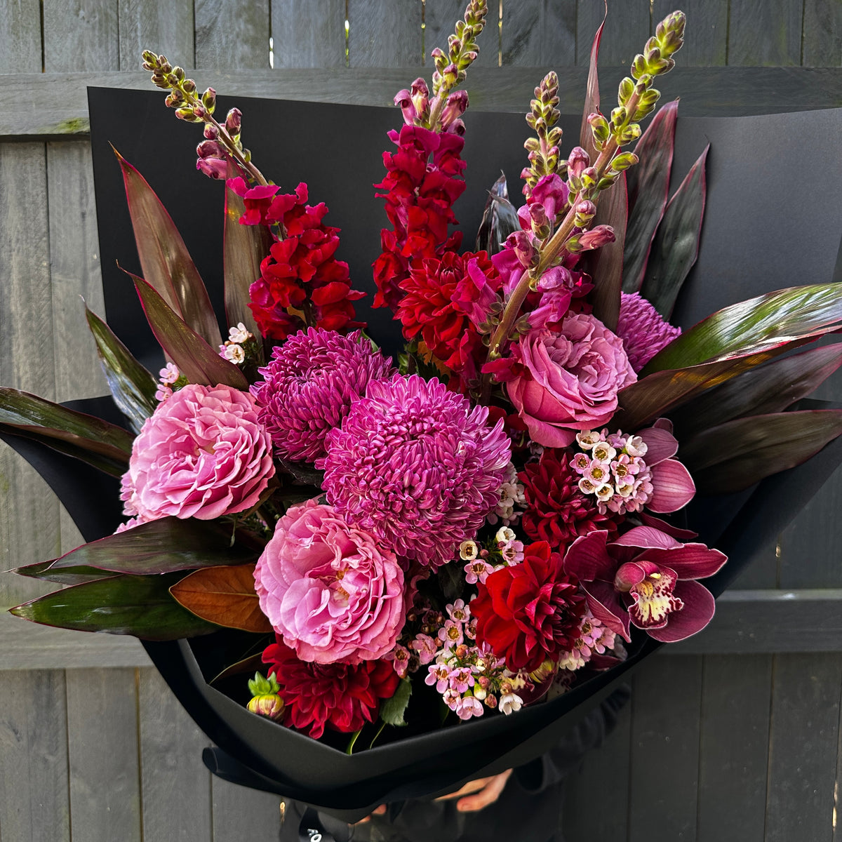Introducing the "September Mood" arrangement by Flowers on Norton St: a vivid bouquet showcasing an exquisite blend of pink, red, and purple roses, snapdragons, dahlias, and chrysanthemums. This stunning floral display is elegantly wrapped in black paper and beautifully contrasted against a wooden fence backdrop.
