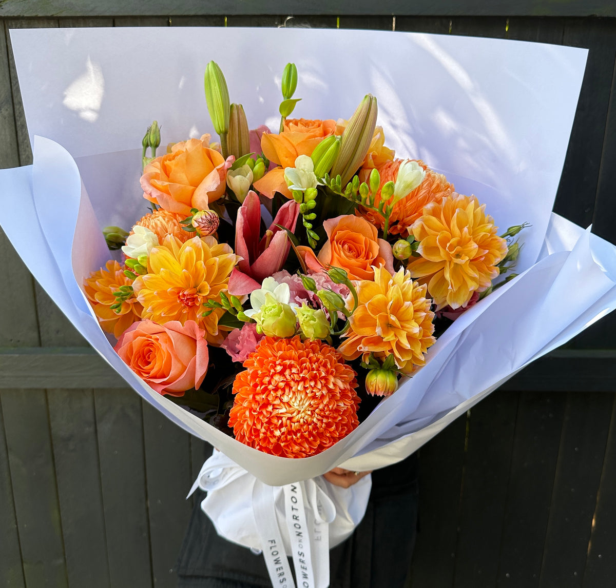 The Spring Picnic bouquet by Flowers on Norton St features a vibrant arrangement of orange and peach roses, tiger lilies, and chrysanthemums with green buds wrapped in white paper, all set against a dark wooden background.