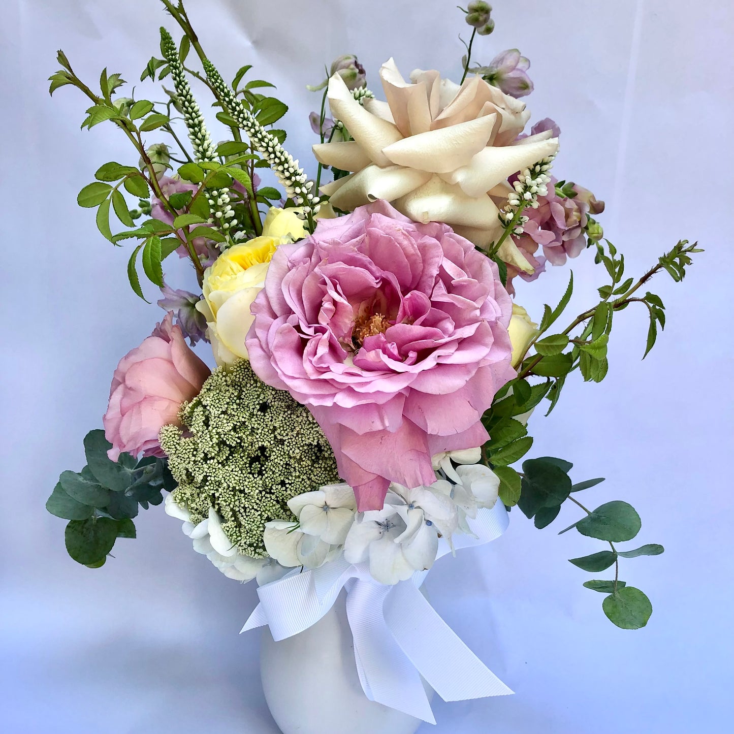 A bouquet in a Flowers on Norton St's Mini Fairy Garden Vase, showcasing hand-flexed pink, white, and yellow roses complemented by seasonal foliage and small white flowers, elegantly tied with a white ribbon.