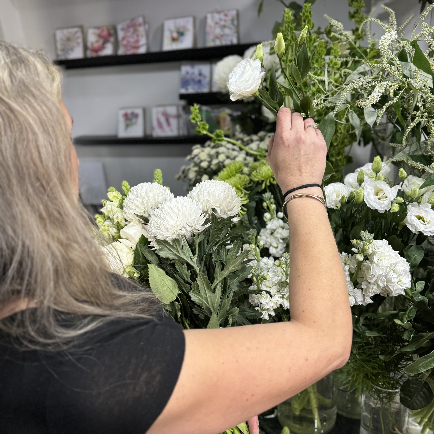 At Flowers on Norton St, a long-haired individual arranges the Florist Choice - White Bouquet with roses and chrysanthemums. Various bouquets and greeting cards in the background showcase affordable flower delivery options in Sydney.