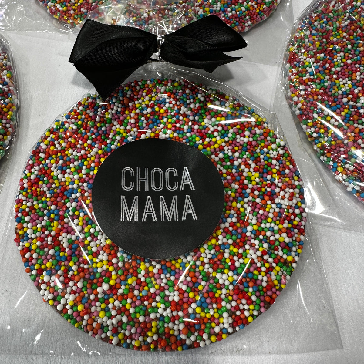 A giant milk chocolate disc with colorful sprinkles is wrapped in clear plastic, tied with a black ribbon. The label reads "Flowers on Norton St | CHOCAMAMA," inviting both kids and adults to enjoy its sweet charm.