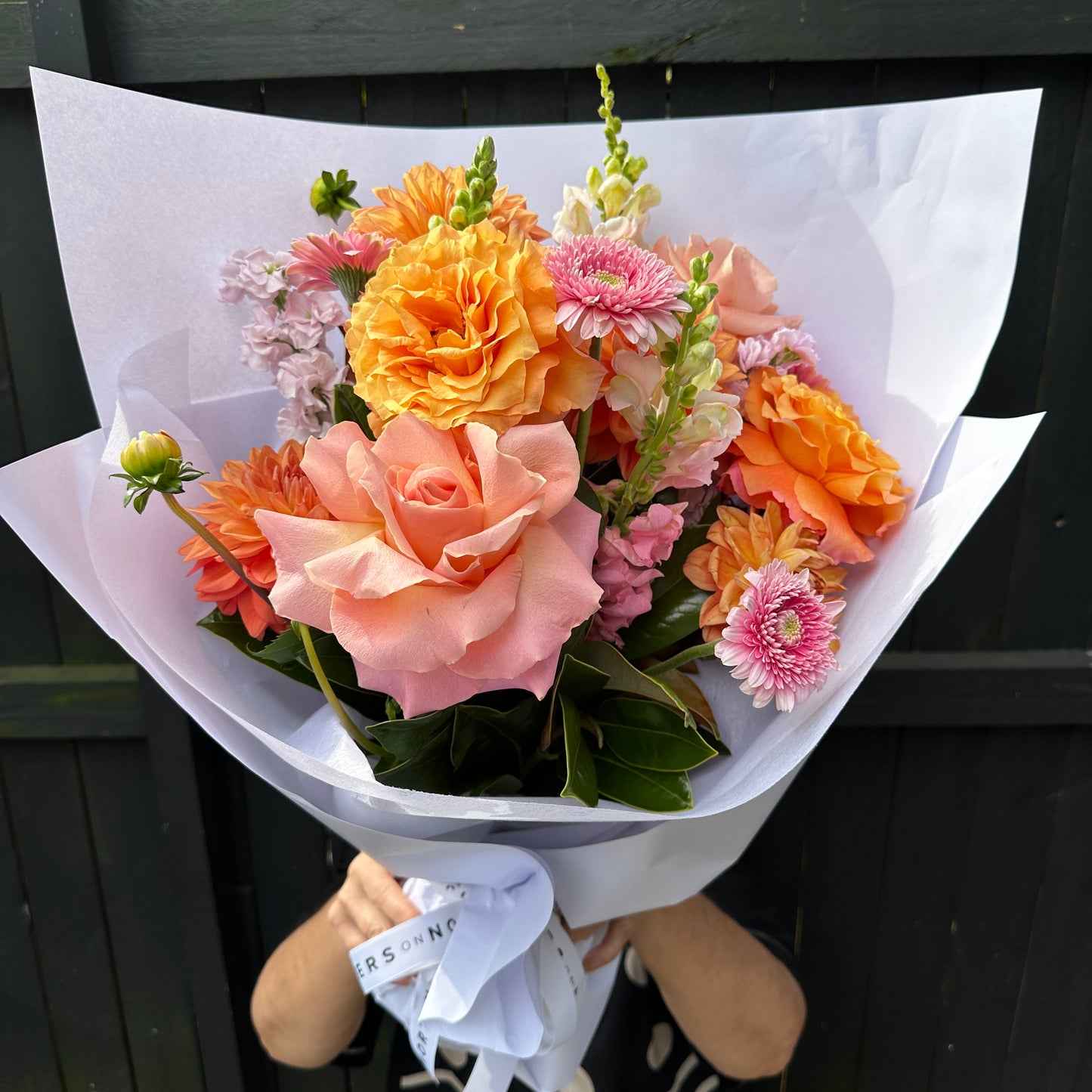 The Starburst Mixed Flower Bouquet by Flowers on Norton St features a vibrant mix of peach and orange roses, pink gerbera daisies, and other seasonal blooms wrapped in white paper, shown against a dark wooden fence for a playful surprise.