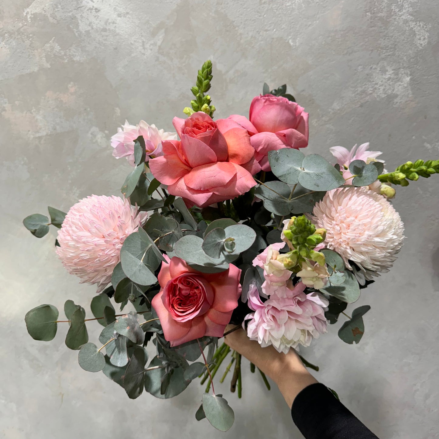 A person holding the "Pretty in Pink Mixed Flowers" bouquet from Flowers on Norton St, featuring pink roses, pale pink chrysanthemums, and eucalyptus leaves against a textured gray background.