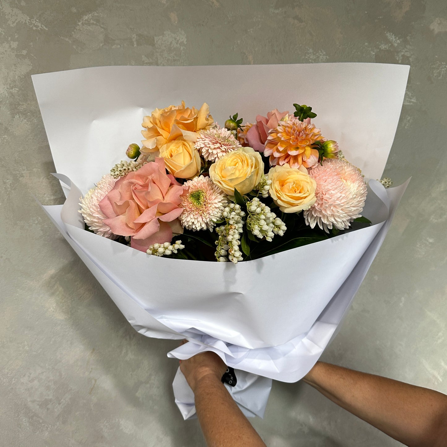 A person holding **The $159 Bouquet** by **Flowers on Norton St**, featuring seasonal blooms like peach roses, yellow roses, pink dahlias, white chrysanthemums, and small white buds wrapped in elegant white paper against a gray background.