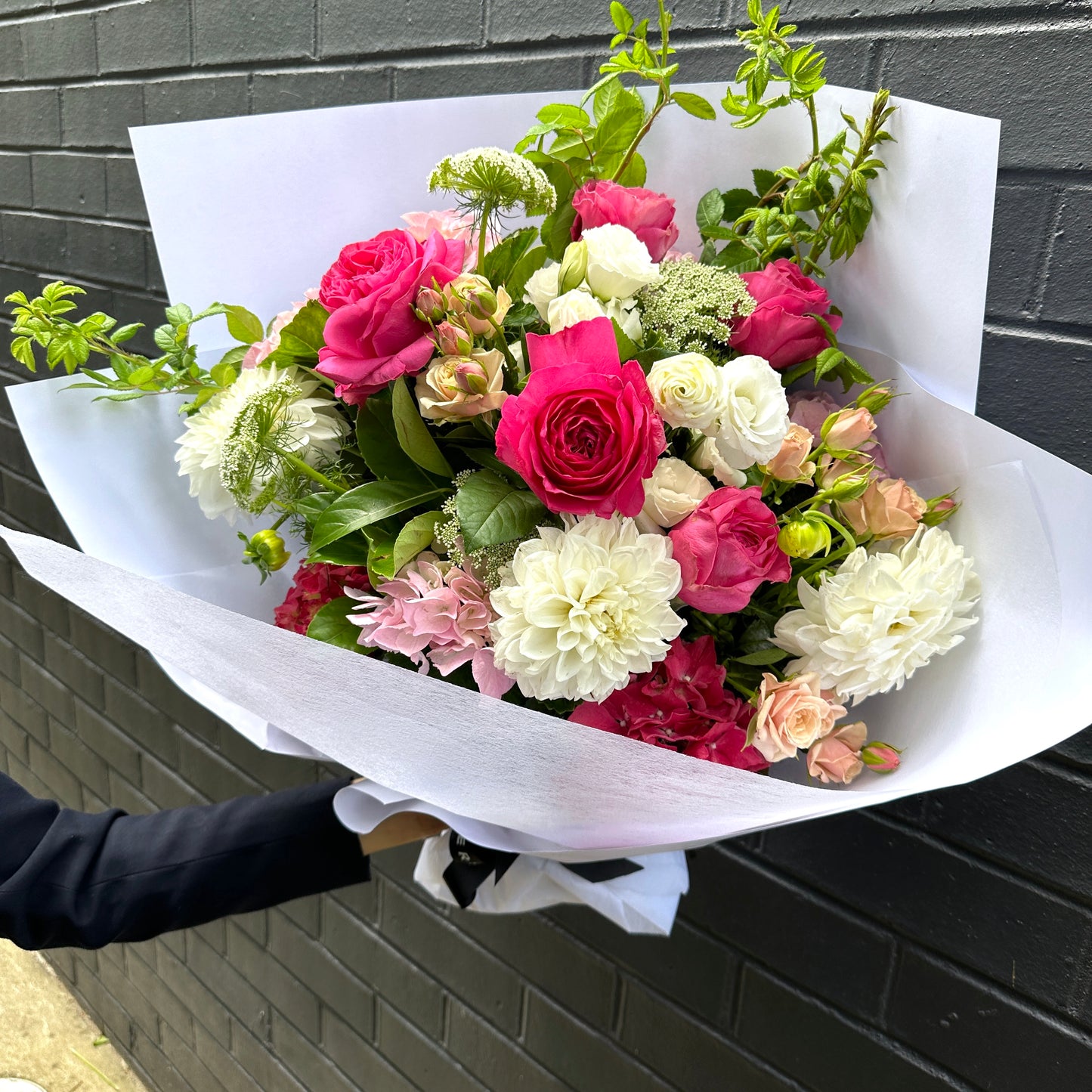 Emma holds a "Summer Garden" bouquet from Flowers on Norton St, with pink and white blooms featuring roses and dahlias, elegantly wrapped in white paper against a dark brick wall.