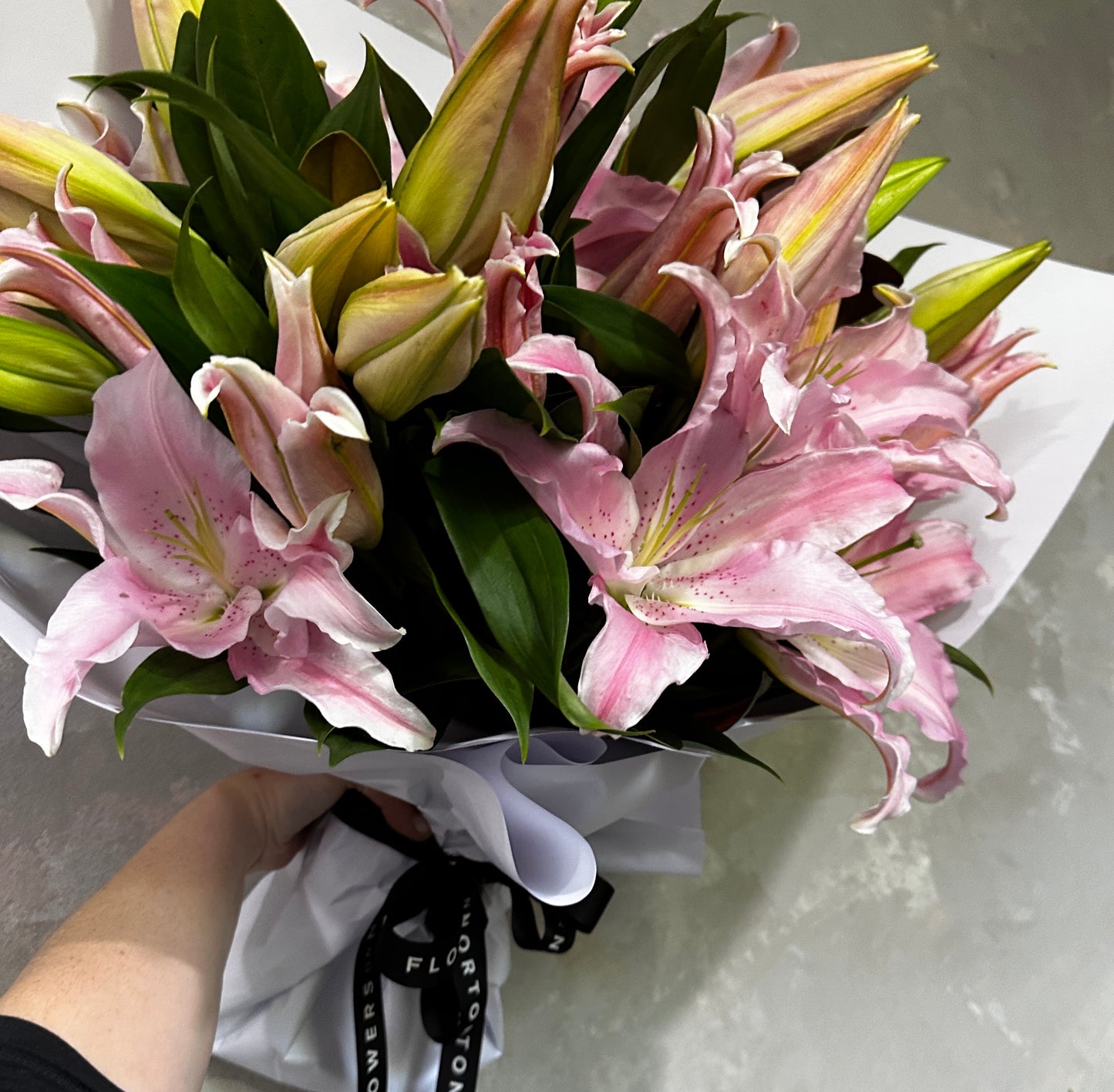 In a person's hand, a bouquet of Casablanca Lily from FlowersonNortonSt, with lush green leaves, is elegantly displayed. The long-lasting pink Oriental lilies are partially wrapped in white paper and secured with a black ribbon, set against a textured gray background that emphasizes their elegance.