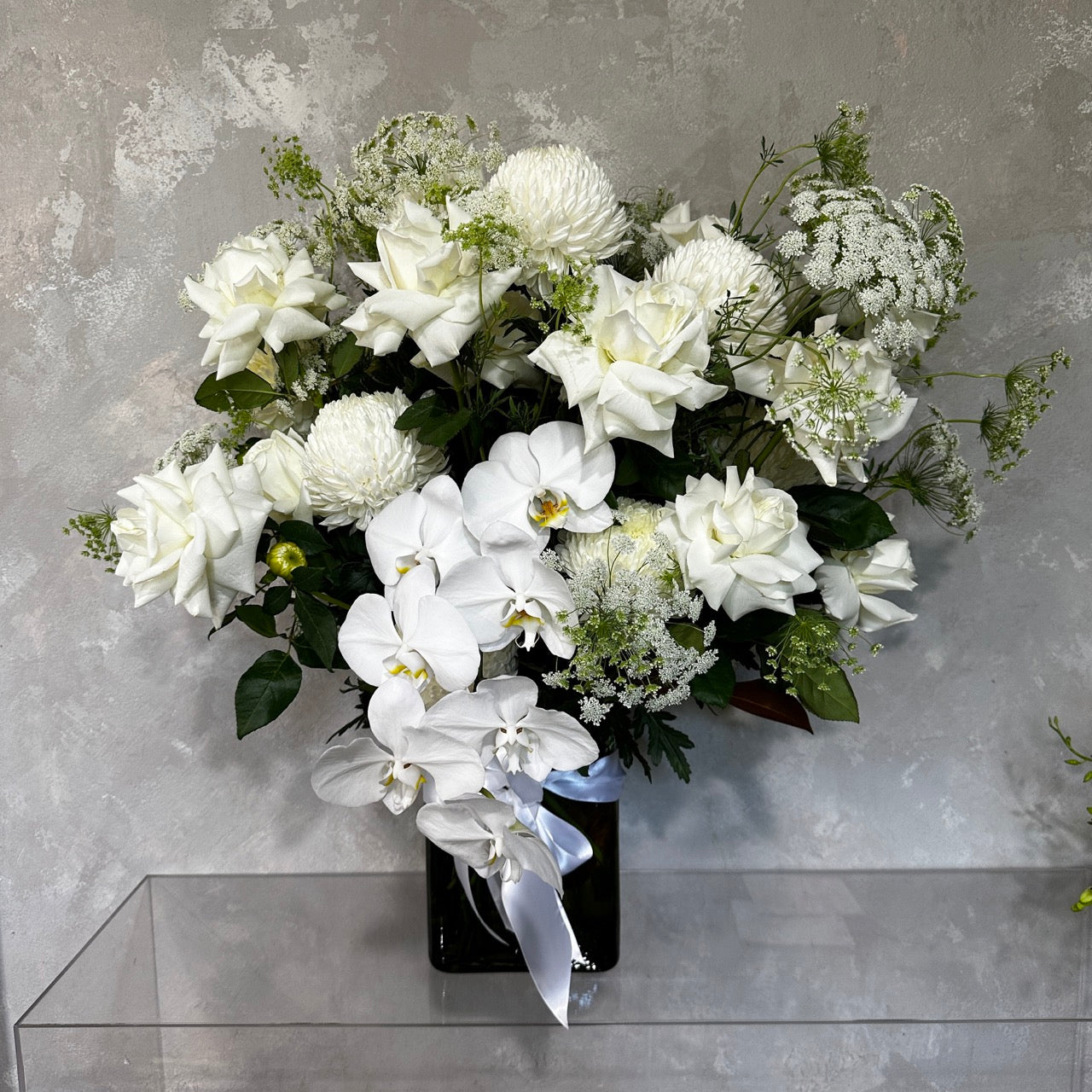 A bouquet of white roses, orchids, and chrysanthemums beautifully arranged with greenery in the elegant Ivory Bliss Mixed Flower Vase from Flowers on Norton St, set against a textured gray background.