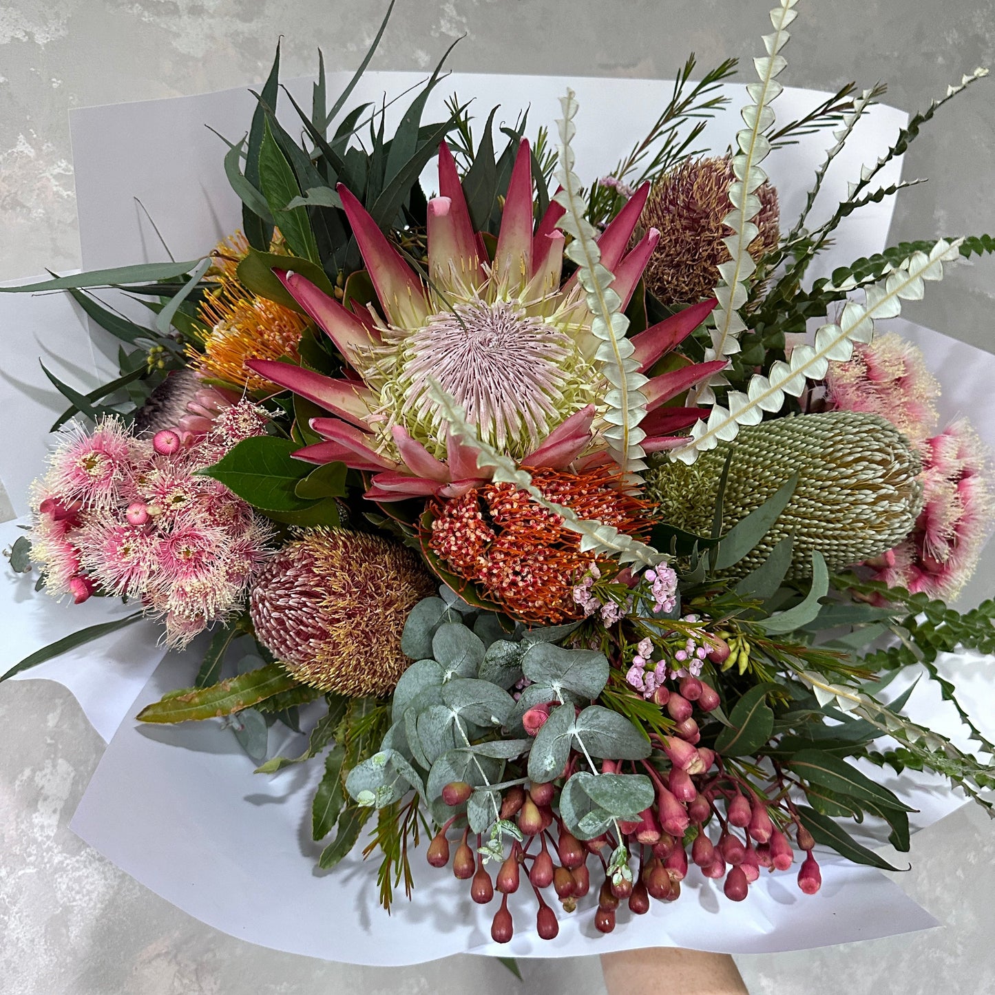 Introducing the "Native Wildflowers" bouquet by Flowers on Norton St: a vibrant arrangement of native protea flowers in stunning shades of pink, red, and orange. This exquisite bouquet is complemented with mixed greenery and small berries, all elegantly wrapped in white paper for a striking contrast with the colorful blooms.