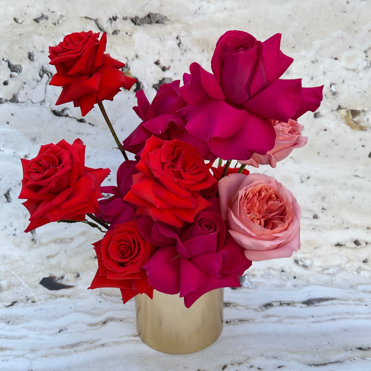 The Veuve Surprise by Flowers on Norton St showcases a vibrant bouquet of roses, featuring deep red, magenta, and light pink blooms nestled in a gold pot. Set against a marbled background, this arrangement highlights the varied textures and rich colors of the flowers.