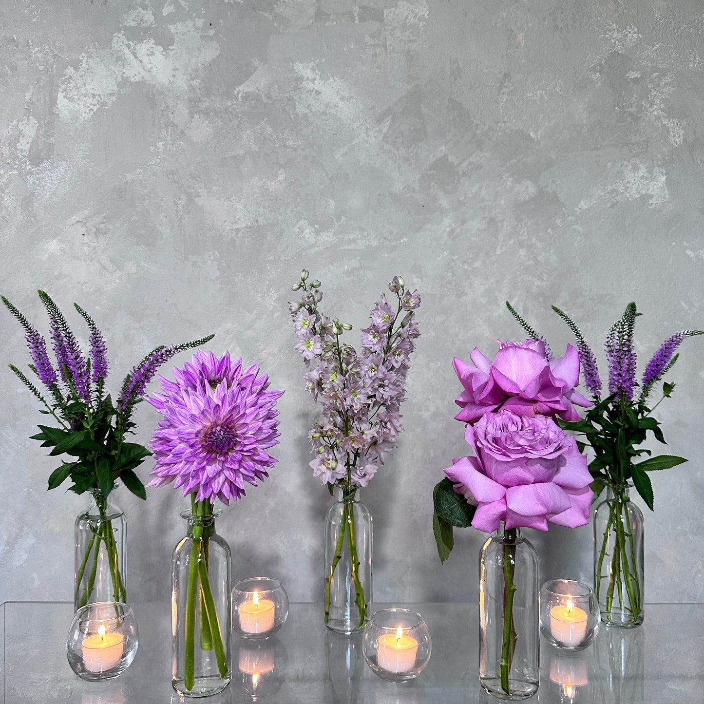 Five elegant glass vases from the Flowers on Norton St "Style the Table Kit - Mother's Day Edition" are artfully arranged with fresh dahlias, roses, and veronicas on a glass surface. Small lit candles flicker gently between the vases, creating a serene ambiance perfect for Mother's Day against a textured gray wall.