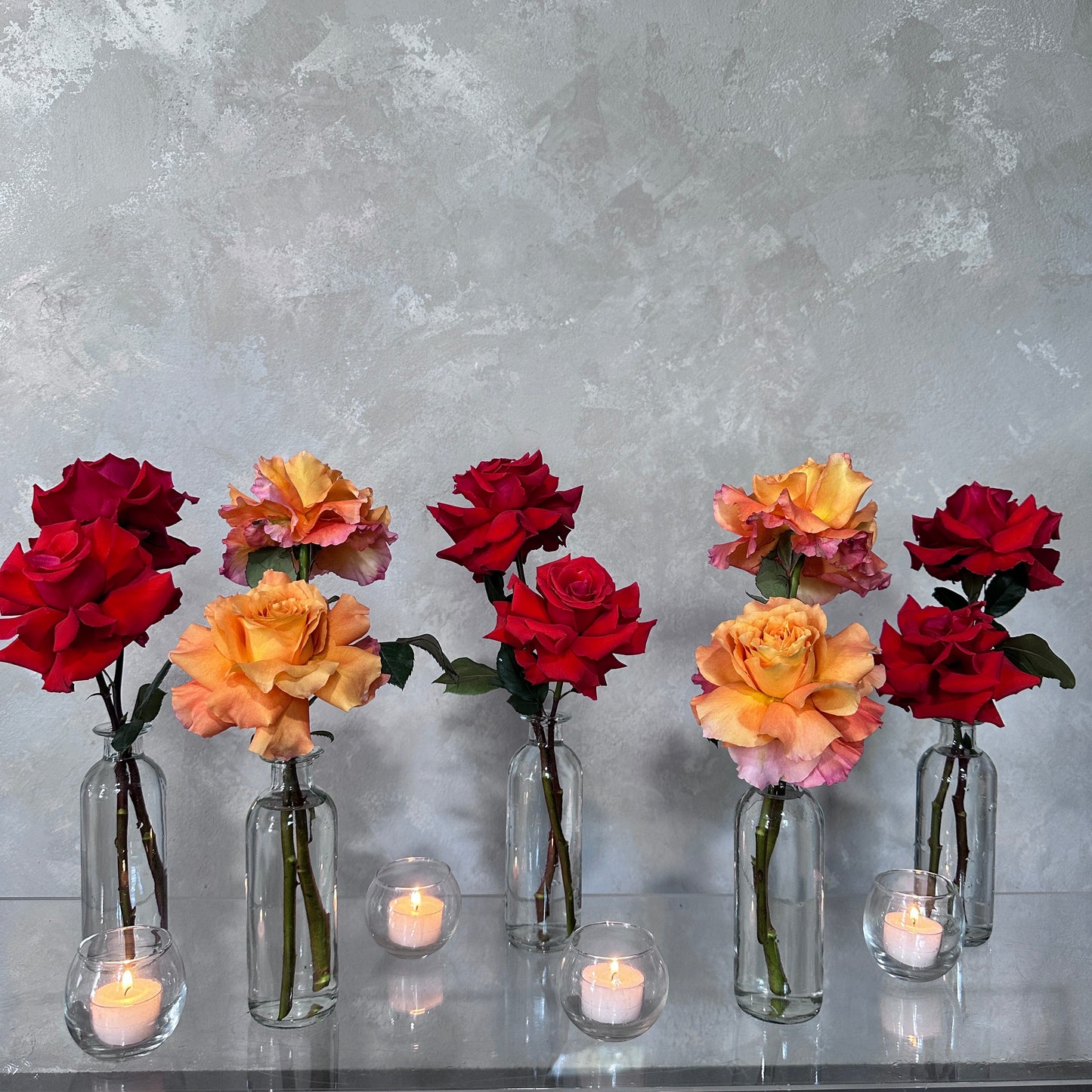 The Style the Table Kit - Mother's Day Edition by Flowers on Norton St features a row of glass vases showcasing fresh florals, including stunning red and orange roses set against a textured gray wall. Small tealight candles in elegant glass holders are stylishly placed in front, enhancing the warm and sophisticated ambiance perfect for Mother's Day celebrations.