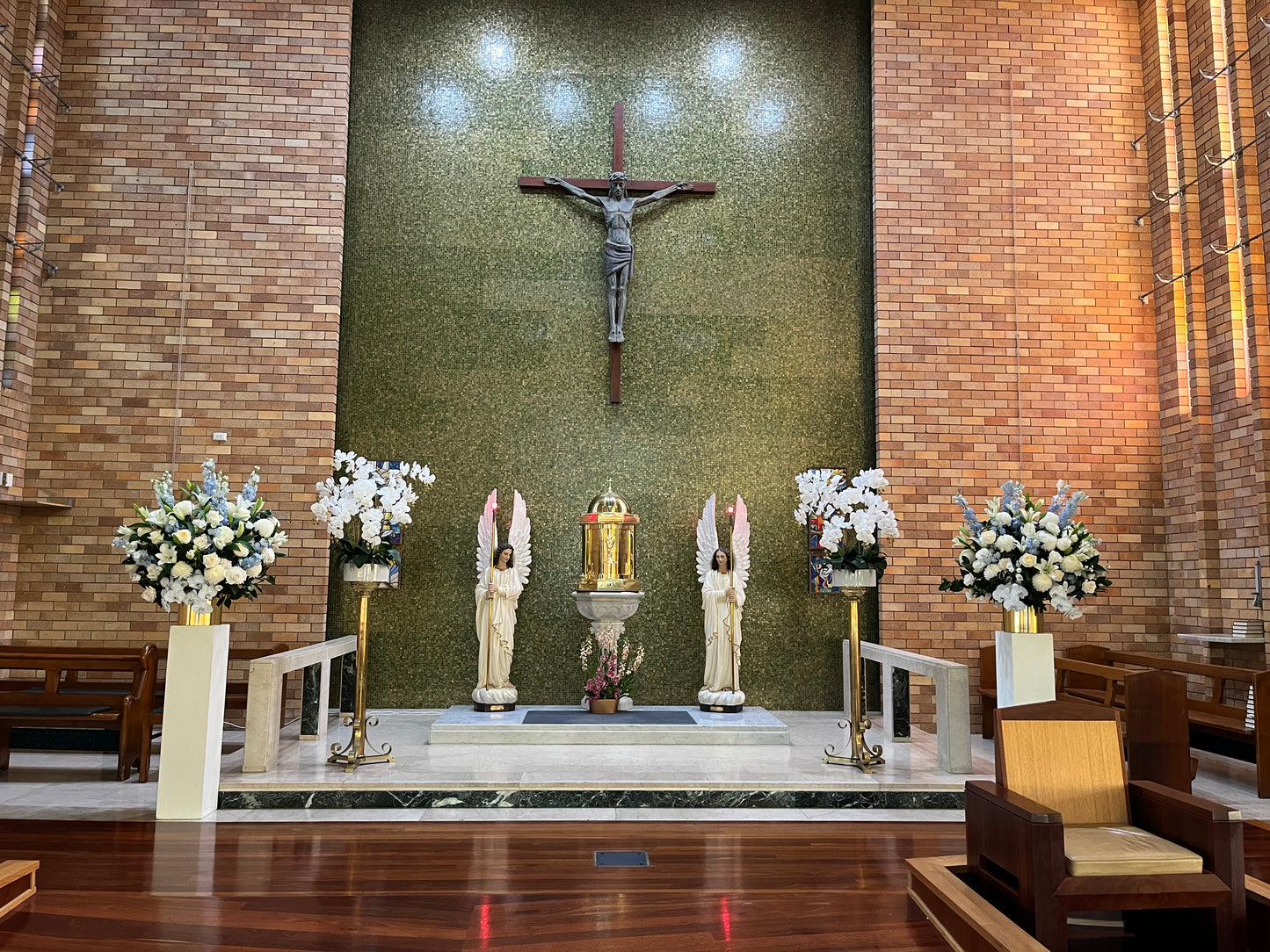 The church altar showcases a prominent crucifix on a green textured wall, accompanied by two angel sculptures on either side of a central golden tabernacle. Encircling the altar are tall ceramic arrangements from Flowers on Norton St., featuring premium florals in white and pink. These arrangements complement the surrounding brick walls and wooden pews beautifully.