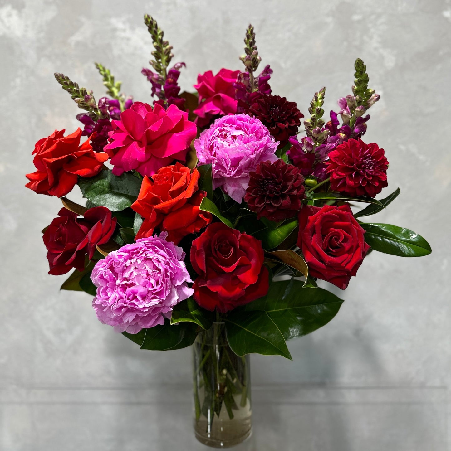 Discover the Romance Mixed Bouquet by Flowers on Norton St: a stunning arrangement of red roses, magenta peonies, deep red dahlias, and lush green leaves in a clear glass vase set against a light gray background.