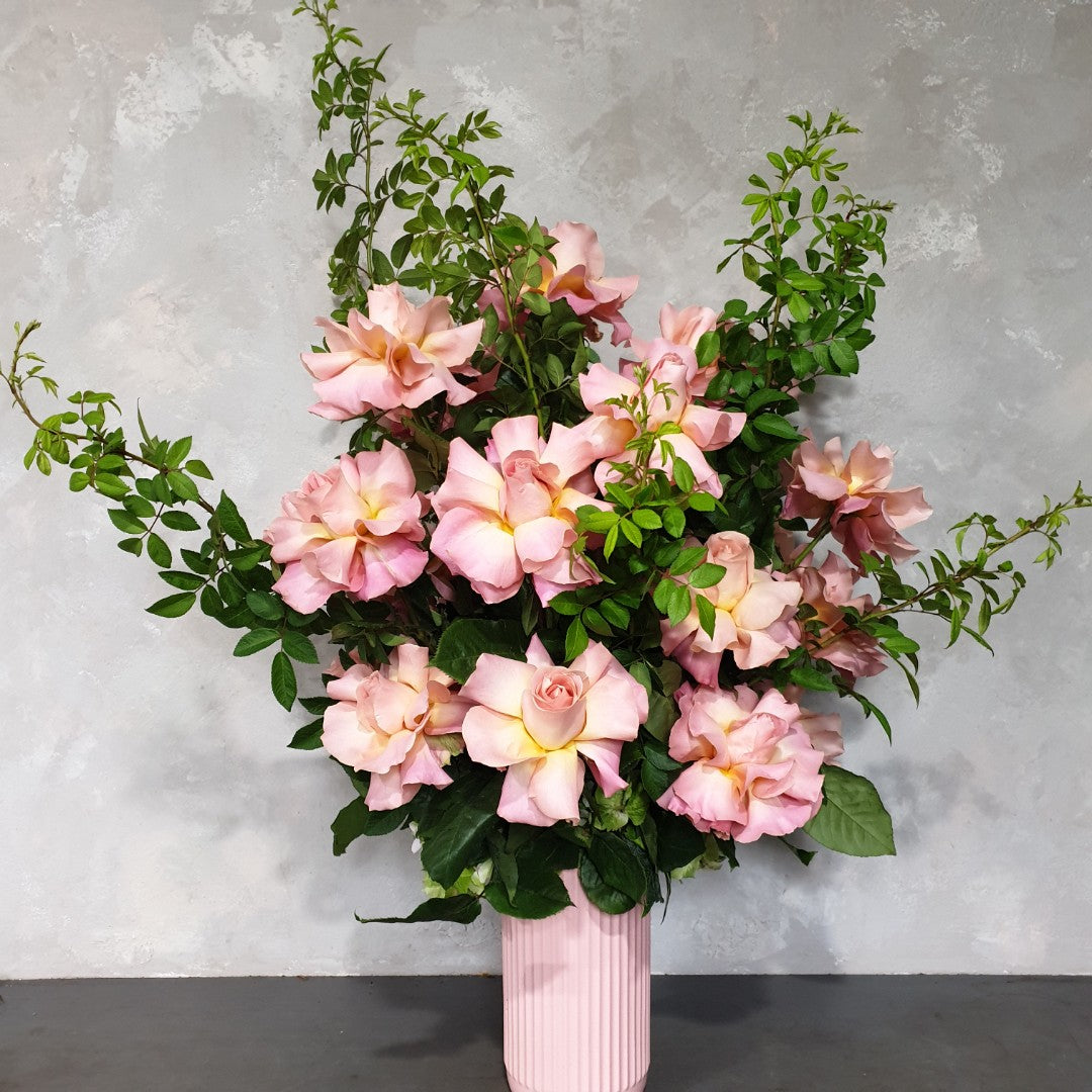 The Ruby Rose Valentine's Vase from Flowers on Norton St showcases Colombian roses with lush green foliage, set against a light gray textured background for an elegant and romantic touch to any setting.