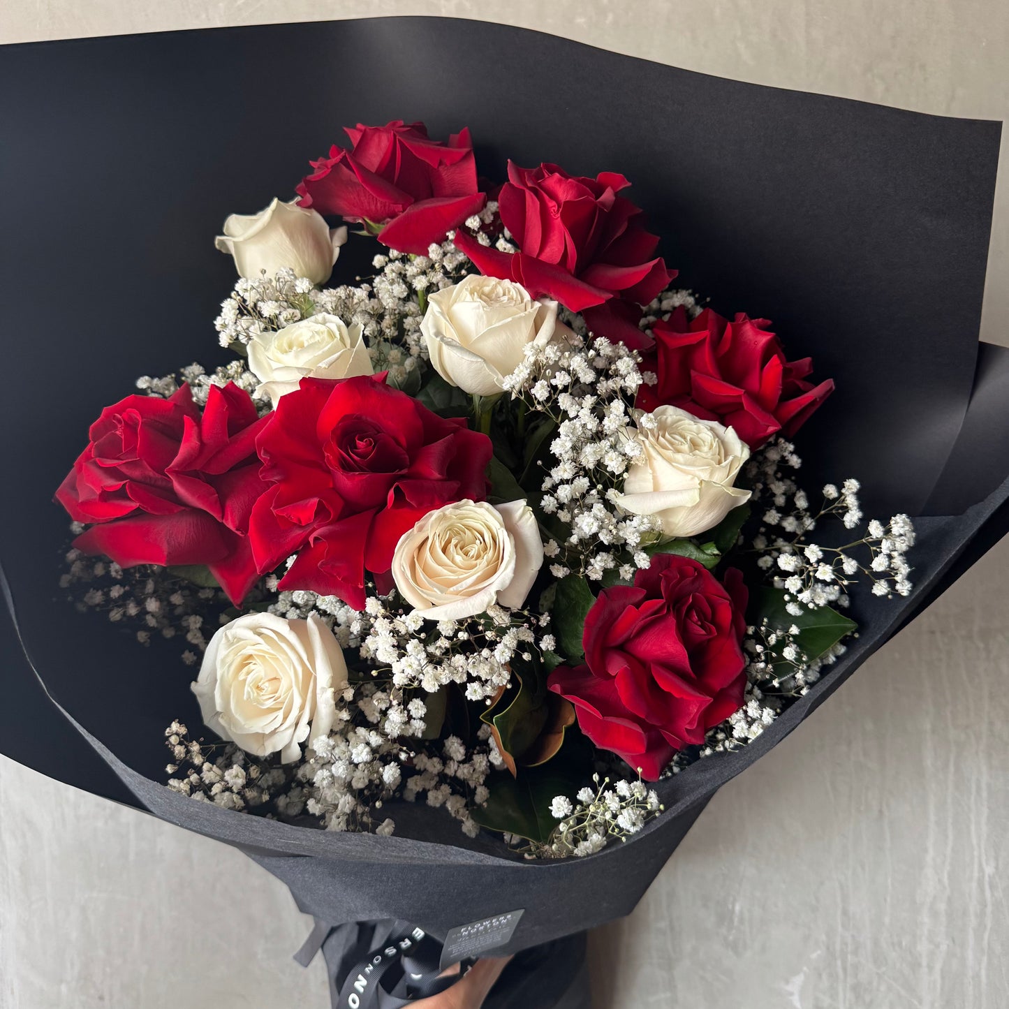 The Bud + Bloom Rose Bouquet with Babies Breath by Flowers on Norton St includes 12 hand-flexed red and white roses, intertwined with baby's breath, and elegantly wrapped in sleek black paper.